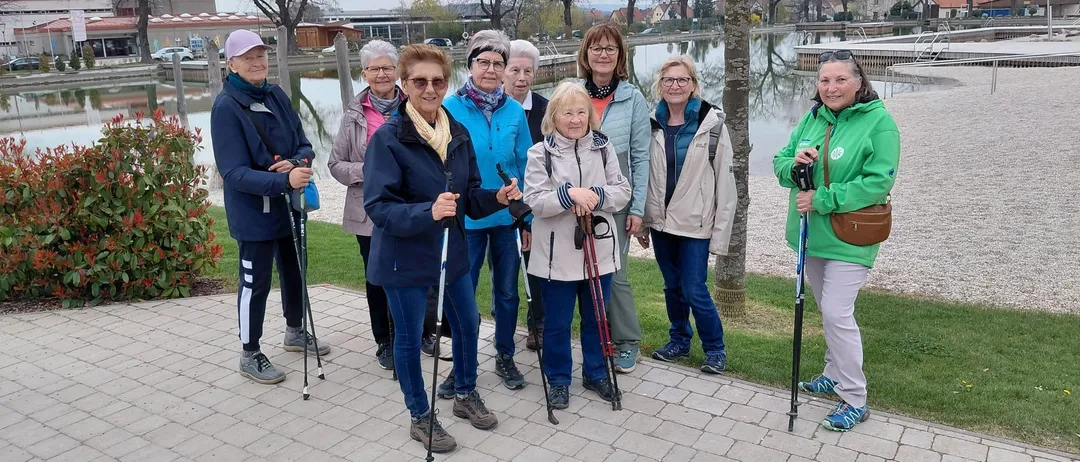 Eine Gruppe älterer Frauen mit Wanderstöcken posiert für ein Foto am See mit Bäumen und Häusern im Hintergrund.
