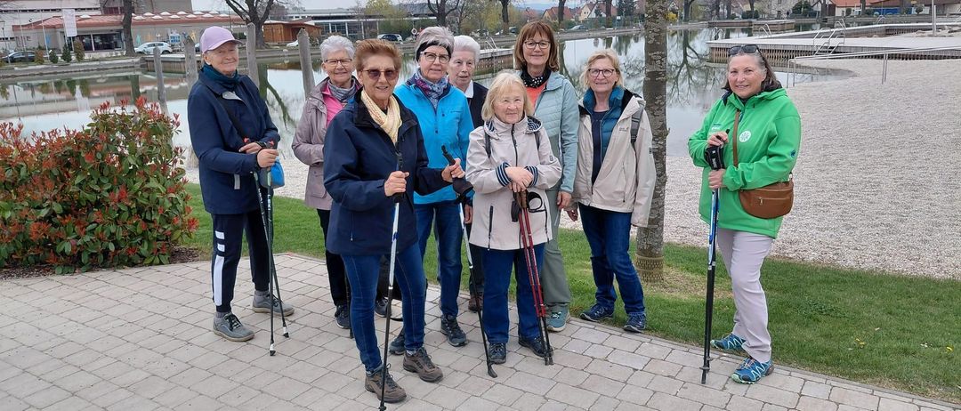 Eine Gruppe älterer Frauen mit Wanderstöcken posiert für ein Foto am See mit Bäumen und Häusern im Hintergrund.