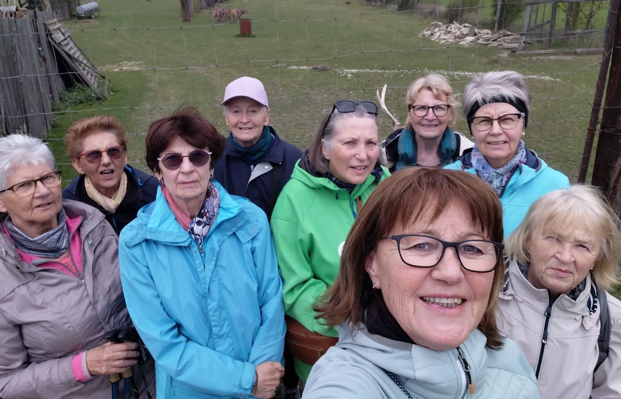 Eine Gruppe von Frauen posiert für ein Foto im Freien, sie tragen Jacken und Sonnenbrillen. Dahinter ist ein Zaun mit Schafen in der Ferne.