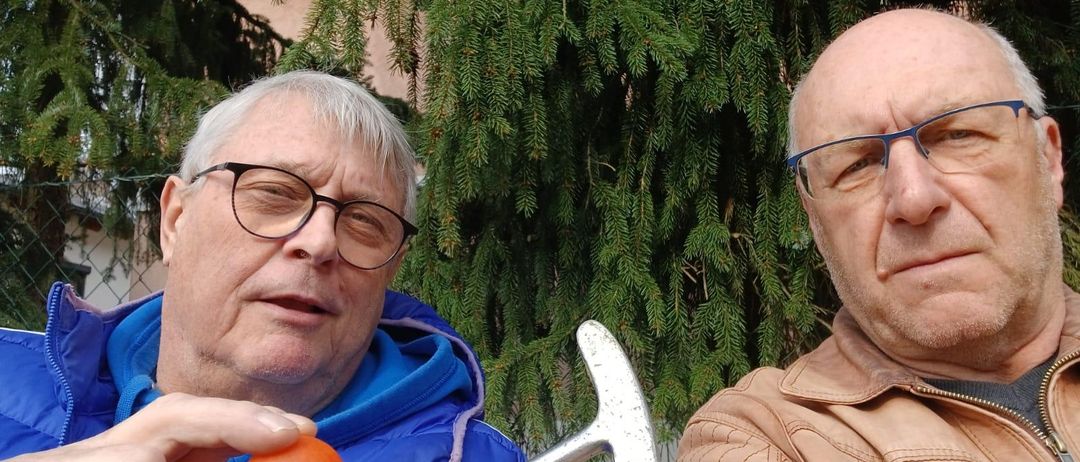 Zwei ältere Männer, einer mit Golfschläger und Ball, posieren vor einer Baumkulisse.