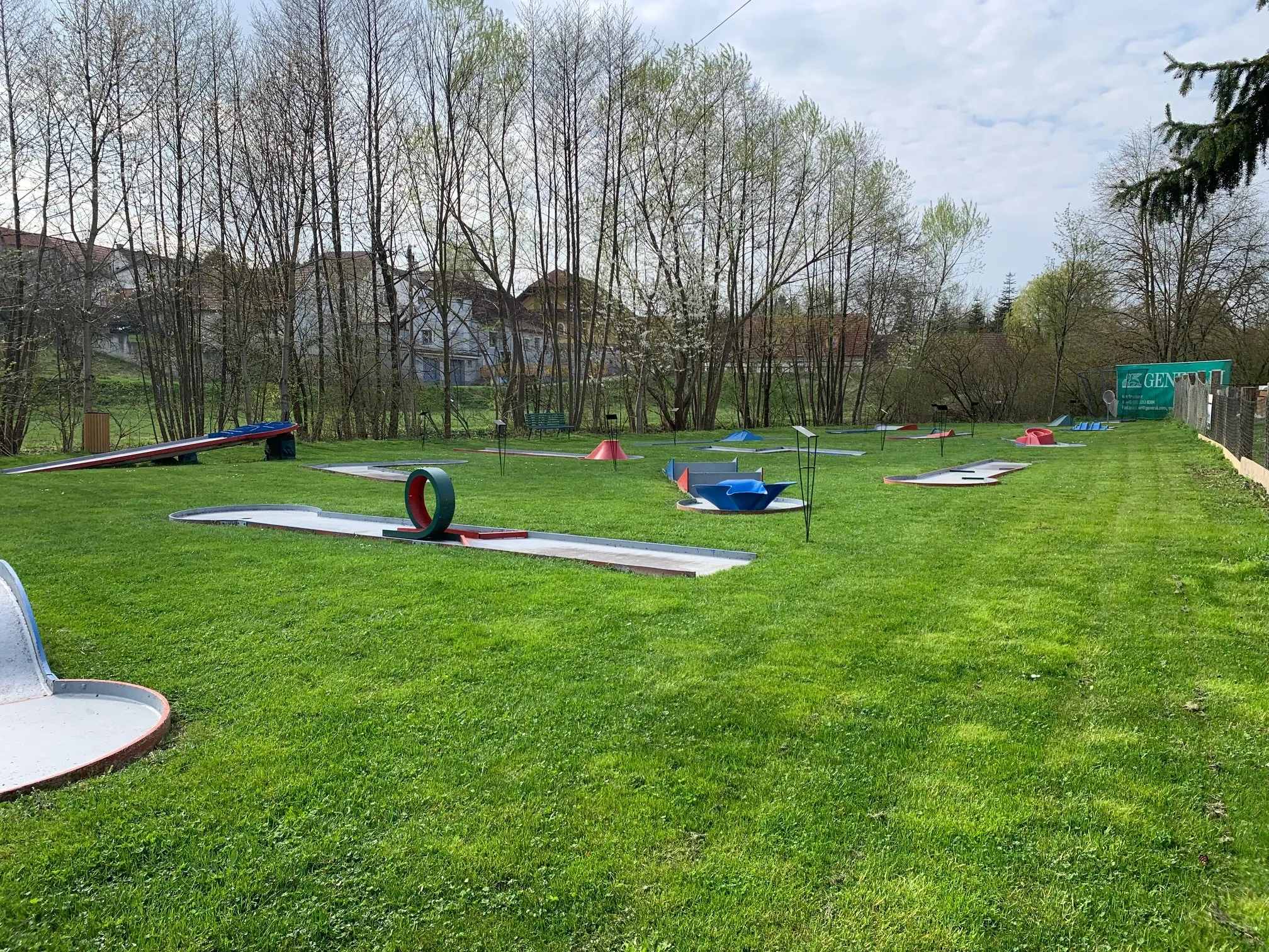 Ein grünes Feld mit verschiedenen bunten Objekten, die darauf verstreut sind. Das Feld ist von Bäumen umgeben und ein Gebäude ist im Hintergrund sichtbar.