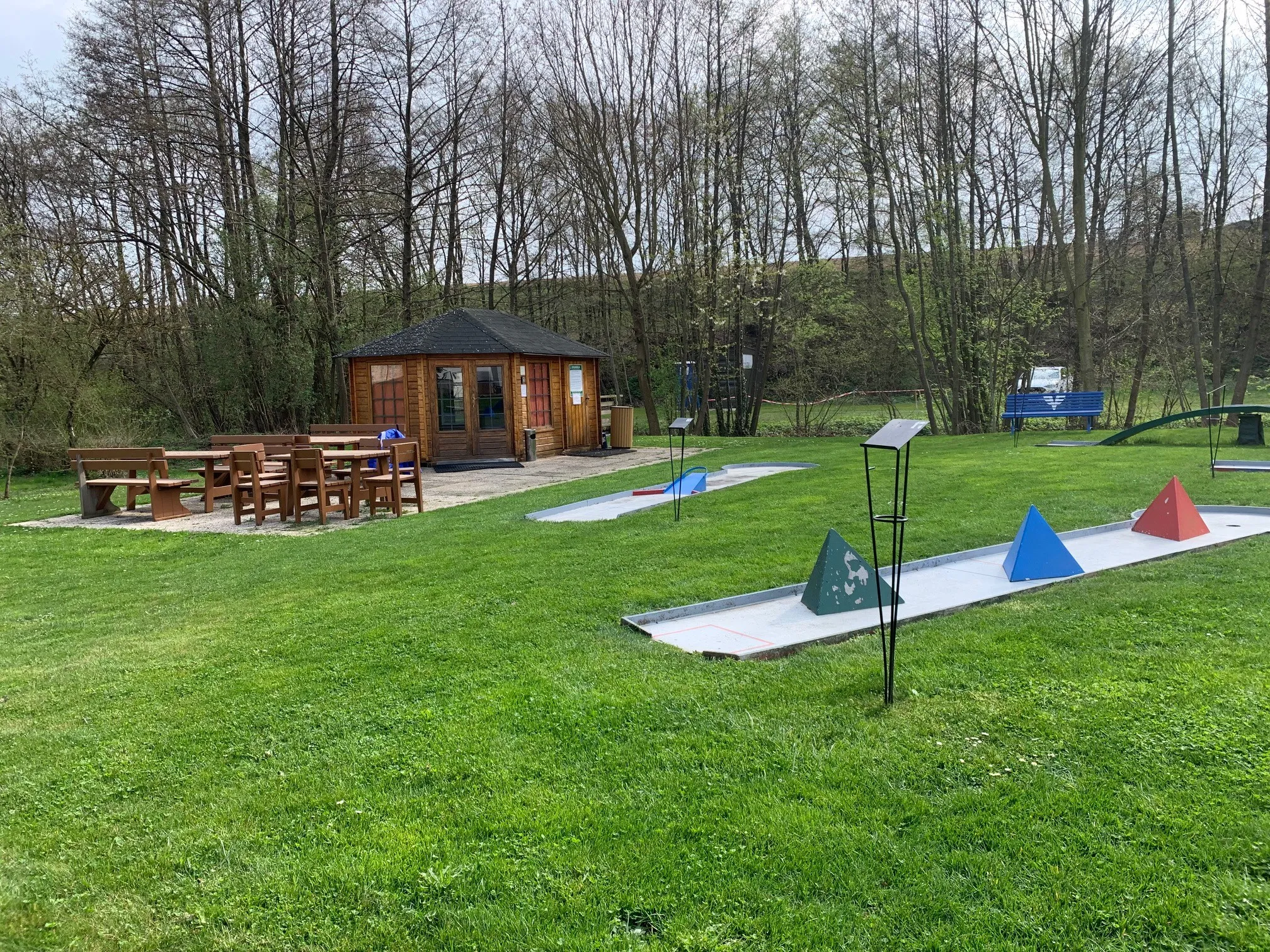 Ein Outdoor-Golfplatz mit zwei Löchern. Es gibt ein Holzhaus und einen Tisch mit Stühlen. Im Hintergrund befinden sich Bäume und eine Bank.