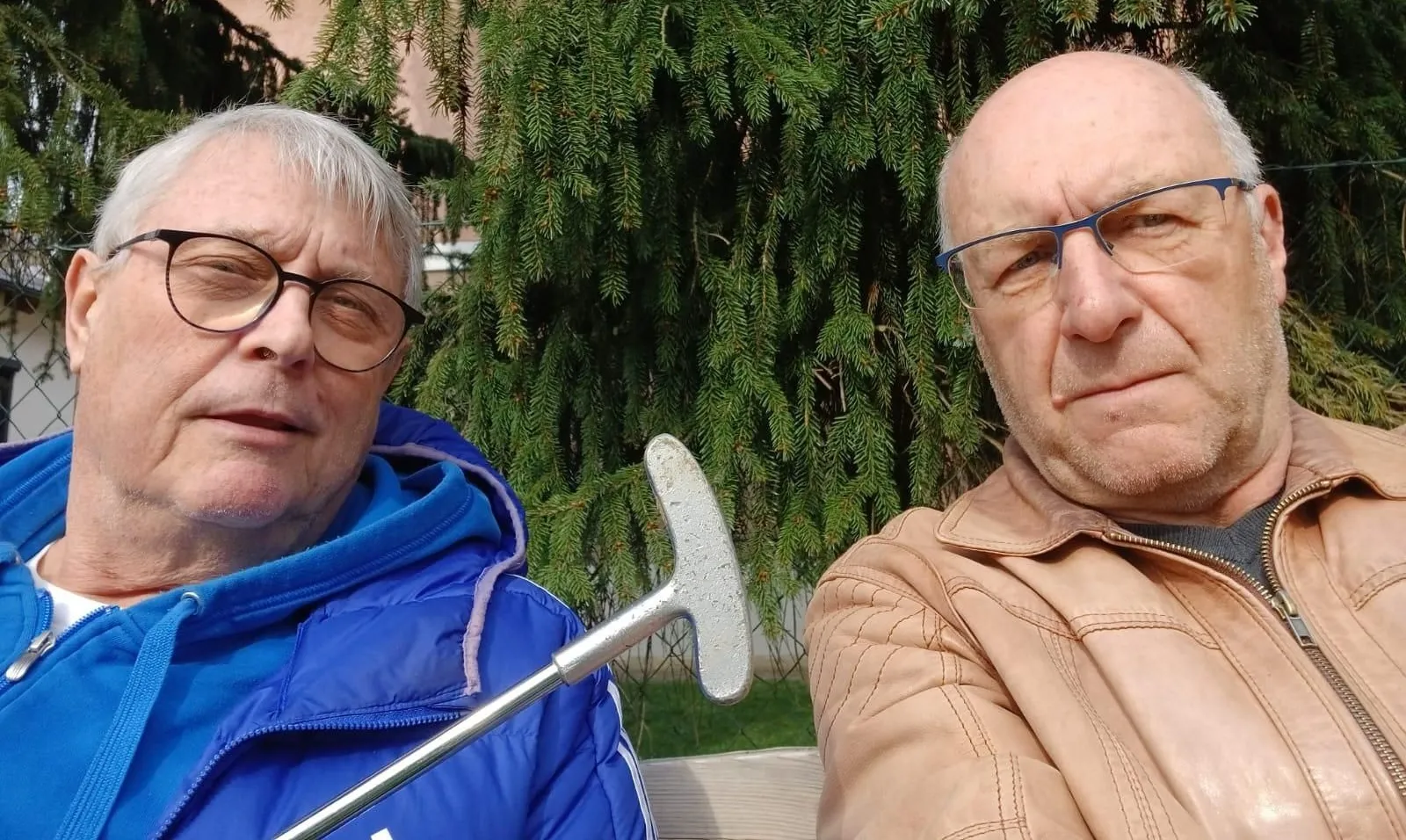 Zwei Männer sitzen auf einer Bank, einer hält einen Golfschläger. Ein Mann trägt eine blaue Jacke und eine Brille, der andere eine beige Jacke und eine Brille. Hinter ihnen steht ein Baum mit dichten grünen Blättern.