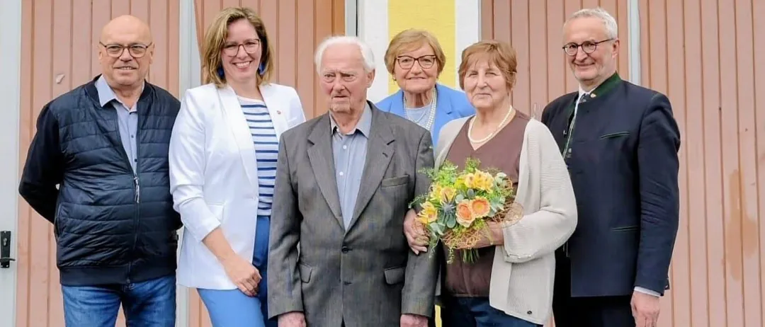 Eine Gruppe von sechs Erwachsenen, drei Männer und drei Frauen, steht vor einem gelben Gebäude mit braunen Türen. Sie alle lächeln für ein Foto.