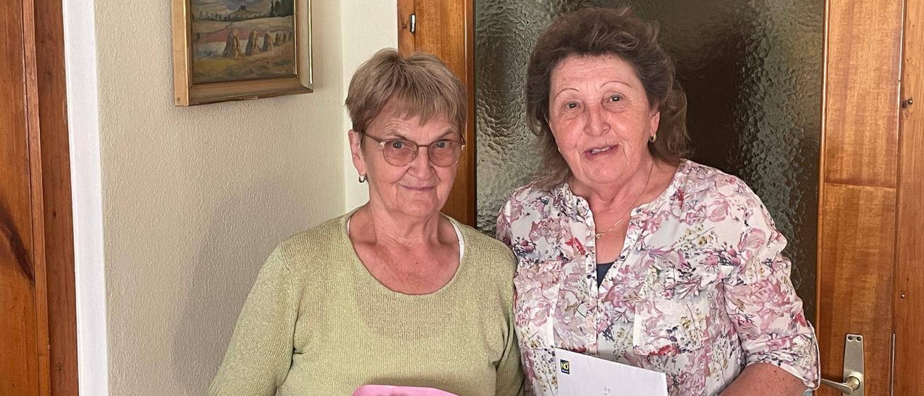 Zwei Frauen stehen nebeneinander, eine hält einen Blumenstrauß und die andere einen Brief. Sie lächeln für ein Foto. Hinter ihnen befindet sich eine Wand mit einem gerahmten Gemälde und einer Glastür.