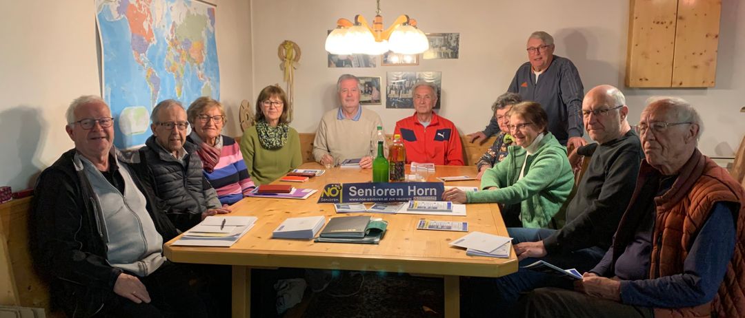 Eine Gruppe älterer Menschen sitzt um einen Tisch, auf dem eine Karte und Flaschen stehen. Das Schild steht für Senioren Horn.