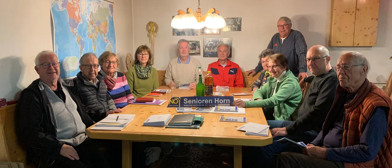 Eine Gruppe älterer Menschen sitzt um einen Tisch, auf dem eine Karte und Flaschen stehen. Das Schild steht für Senioren Horn.