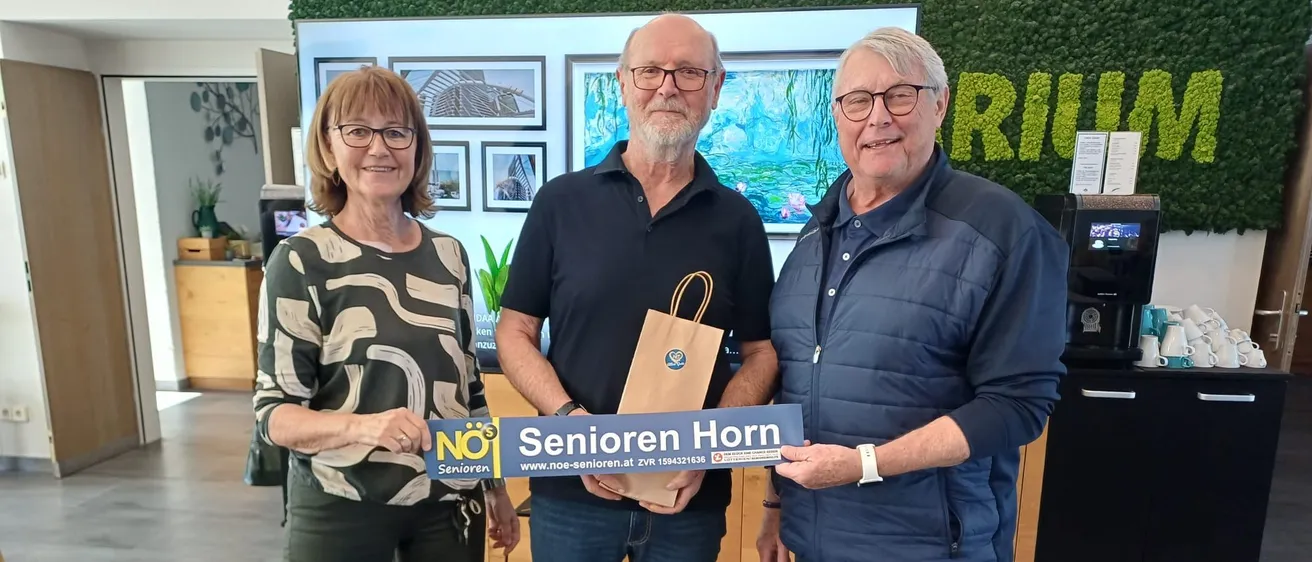 Drei Menschen stehen nebeneinander, lächeln und halten ein blaues Schild mit der Aufschrift 'Senioren Horn'. Einer von ihnen hält eine Papiertüte. Hinter ihnen ist eine Wand mit einer Wandmalerei und Bildern.
