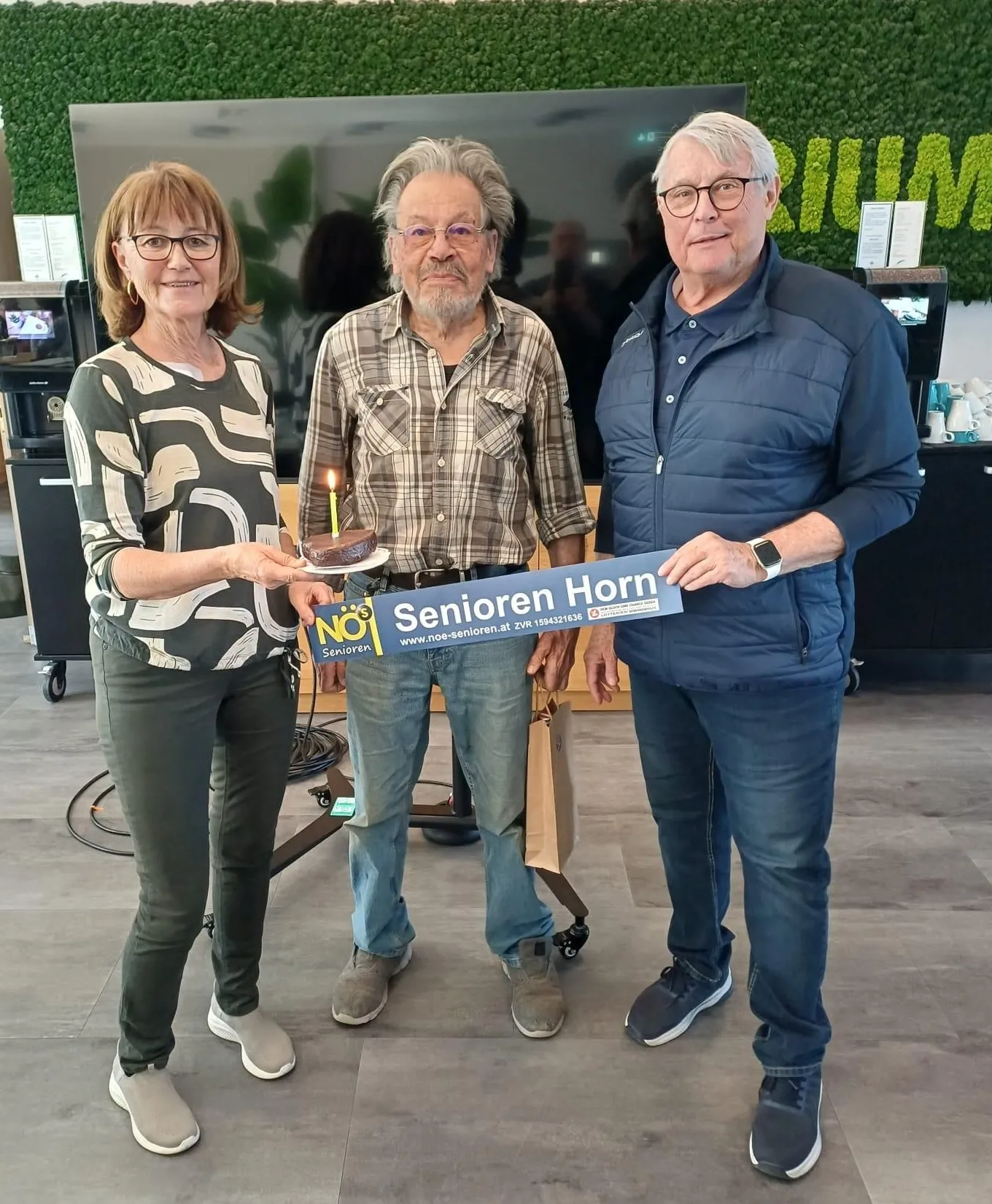 Drei Personen stehen in einem Innenraum und feiern einen Geburtstag. Ein Mann hält ein Banner und einen Kuchen mit einer Kerze. Ein anderer Mann hält einen braunen Papierbeutel. Eine Frau hält einen Kuchen.