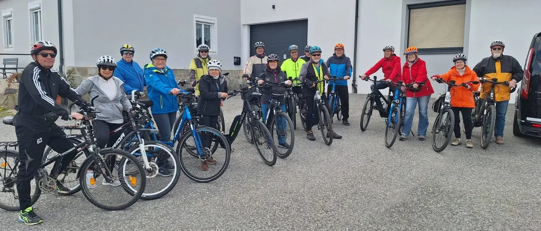 Eine Gruppe von Radfahrern mit Helmen und bunten Jacken steht vor einem weißen Haus mit mehreren Fahrrädern.