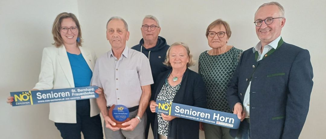 Sechs Personen stehen in einer Reihe, lächeln für ein Foto, jeder hält ein blaues Banner mit NOJ darauf. Zwei Frauen tragen Brillen und eine hält eine Plakette.