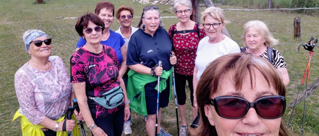 Eine Gruppe von Frauen in Wanderausrüstung steht in einem Feld mit einem Holzpfahl im Hintergrund. Sie alle lächeln und posieren für ein Foto.