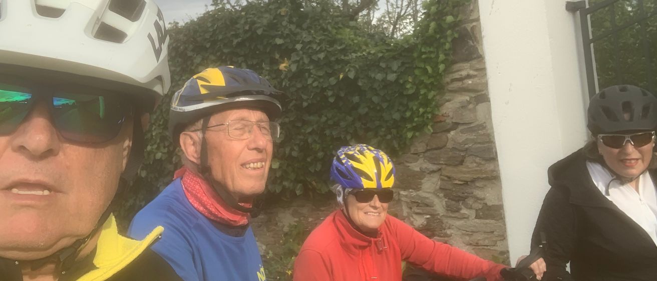 Drei Menschen lächeln und fahren Fahrräder, einer mit roter Jacke und Helm, einer in Blau und eine verschwommene Person in Schwarz. Sie sind von grünen Pflanzen und einer Steinmauer umgeben.