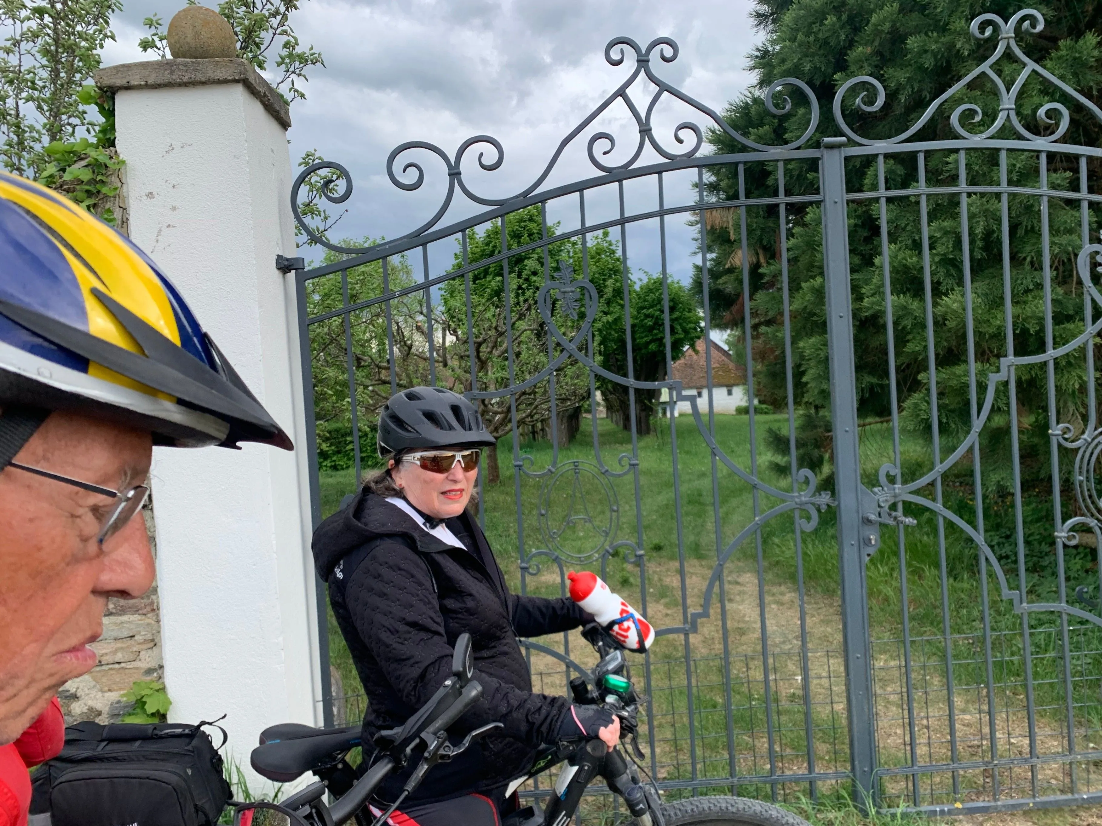 Eine Frau in einer schwarzen Jacke und Helm fährt ein Fahrrad, mit einer Wasserflasche in der Hand, vor einem kunstvollen Tor stehend.