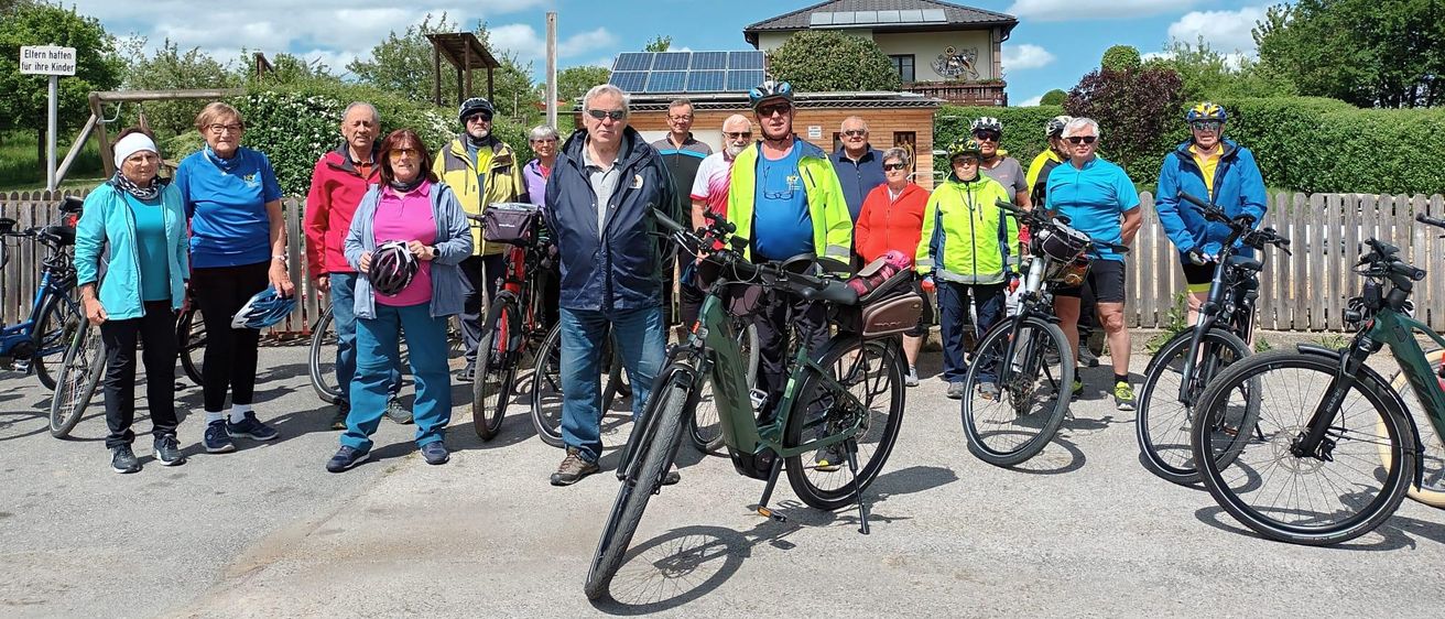 Eine Gruppe von Radfahrern steht an einem sonnigen Tag zusammen, in der Nähe eines Hauses mit Solarmodulen und Grünpflanzen. Sie tragen Helme und Fahrradausrüstung.