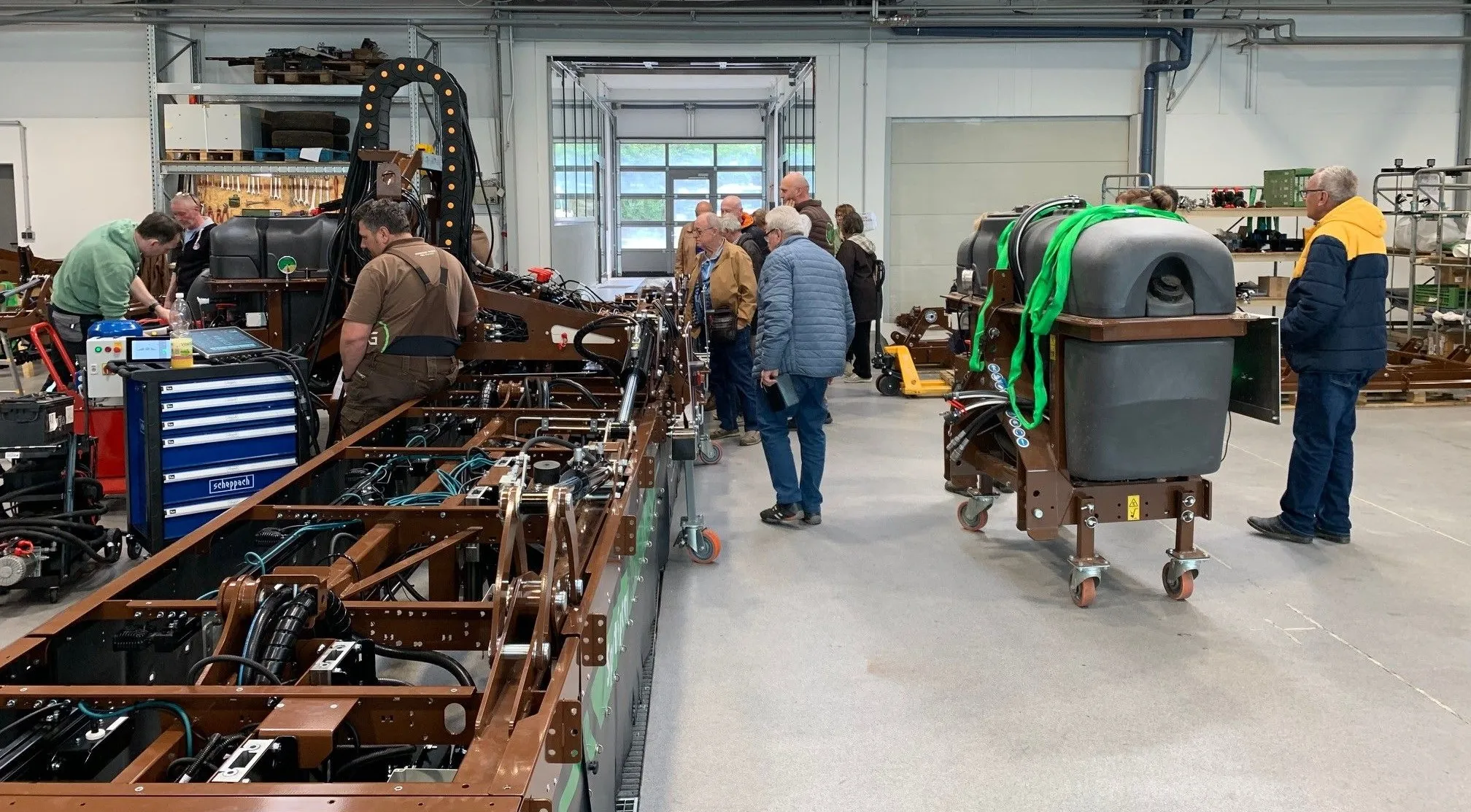 In einer Fabrik untersuchen einige Personen eine große, komplexe Maschine. Arbeiter stellen Teile ein, während andere zuschauen. Geräte sind ordentlich aufgestellt, einige werden auf Karren transportiert.