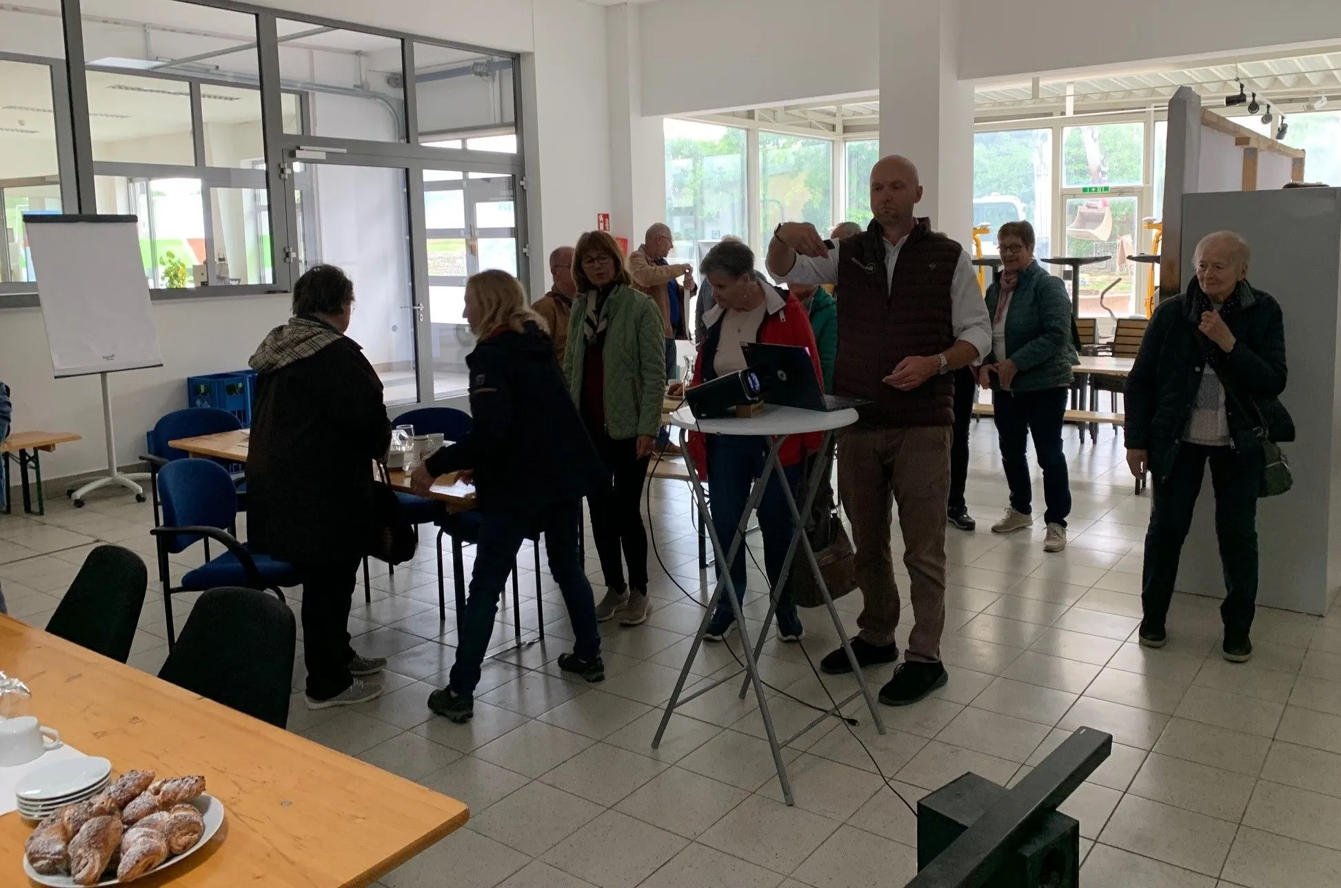 Eine Gruppe von Menschen versammelt sich in einem Raum, in dem ein Mann eine Präsentation mit einem Laptop auf einem Tisch hält. Der Raum ist gut beleuchtet und hat große Fenster.