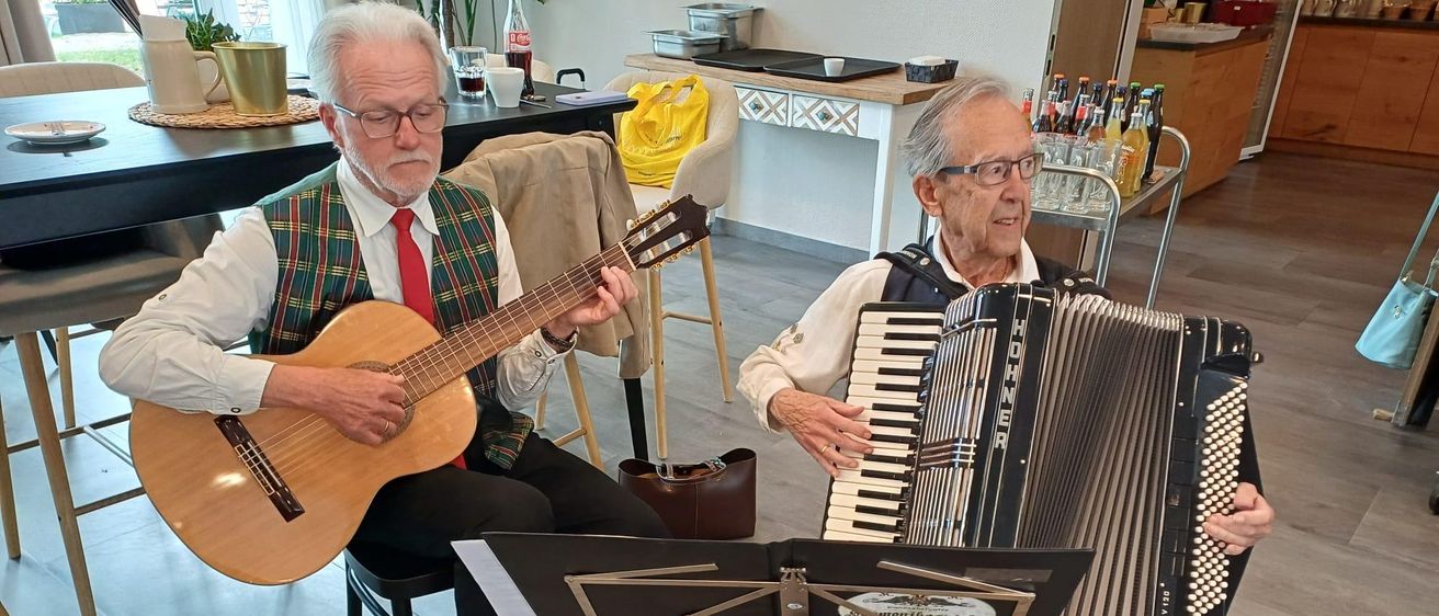 Zwei ältere Männer spielen Instrumente; einer spielt Gitarre, der andere Akkordeon. Sie sitzen auf Stühlen mit Notenständern vor ihnen. Hinter ihnen steht ein Tisch mit Tassen und einem gelben Beutel.