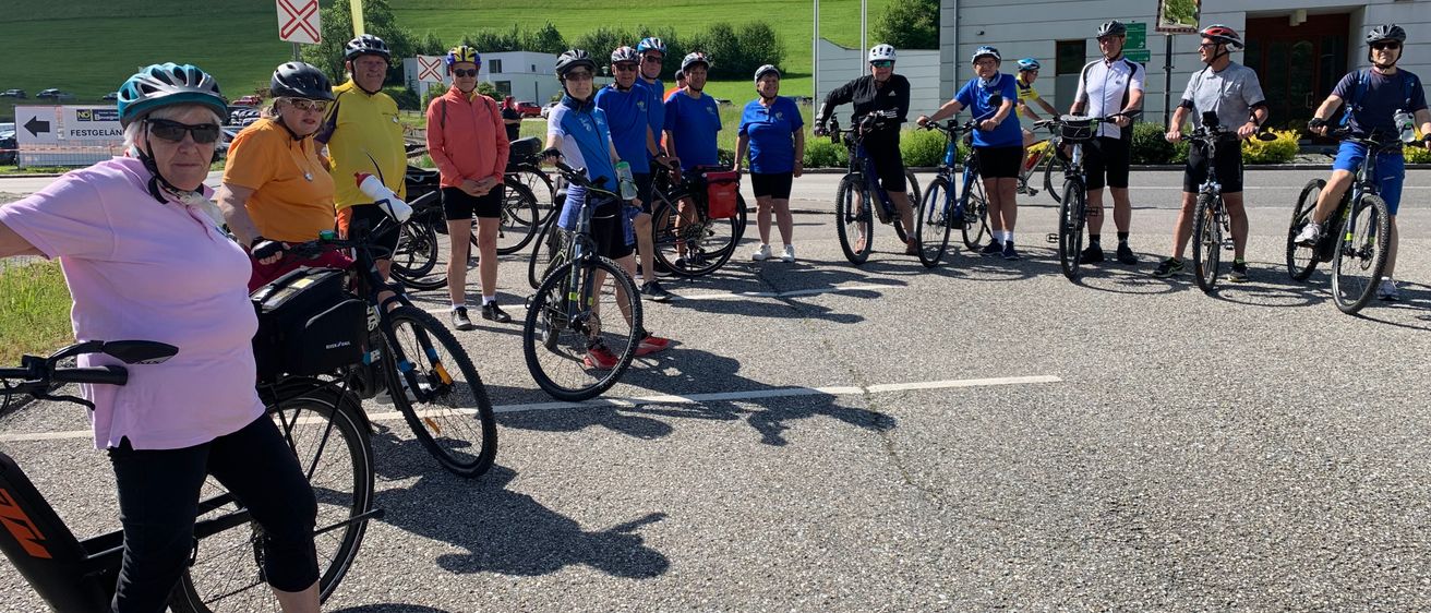 Eine Gruppe von Radfahrern ist auf einem Parkplatz versammelt, mit Fahrrädern in einer Reihe. Sie tragen Helme und einige haben Handschuhe. Im Hintergrund sieht man einen grün bewachsenen Hügel und ein Gebäude.