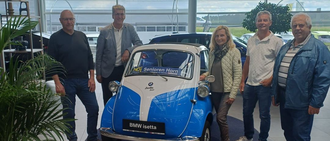 Fünf Personen posieren mit einem blauen BMW Isetta in einem Ausstellungsraum. Das Auto hat 'Senioren Horn' an der Seite geschrieben. Hinter ihnen sind Autos geparkt.