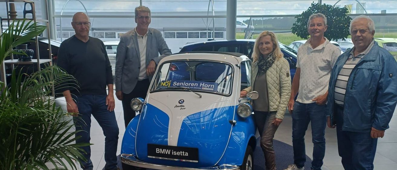 Fünf Personen posieren mit einem blauen BMW Isetta in einem Ausstellungsraum. Das Auto hat 'Senioren Horn' an der Seite geschrieben. Hinter ihnen sind Autos geparkt.