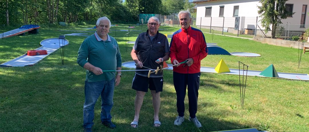 Drei Männer stehen auf einem Golfplatz. Ein Mann hält einen Golfschläger. Die anderen lächeln. Hinter ihnen sind Bäume und ein Haus.