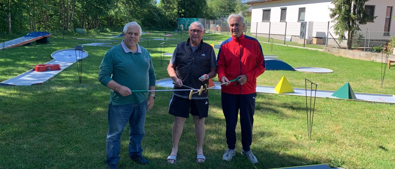 Drei Männer stehen auf einem Golfplatz. Ein Mann hält einen Golfschläger. Die anderen lächeln. Hinter ihnen sind Bäume und ein Haus.