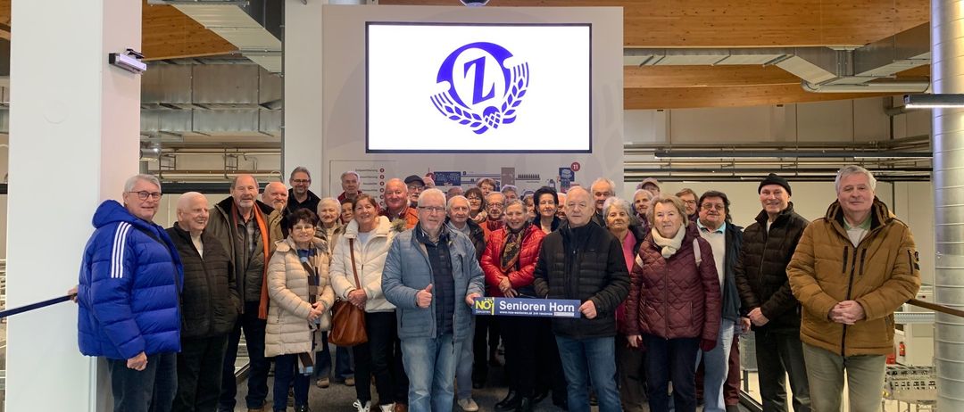 Eine Gruppe von Menschen posiert für ein Foto in einem Saal. Ein großer Bildschirm zeigt das Logo von 'Senioren Horn'. Sie sind warm gekleidet, einige halten Handtaschen.