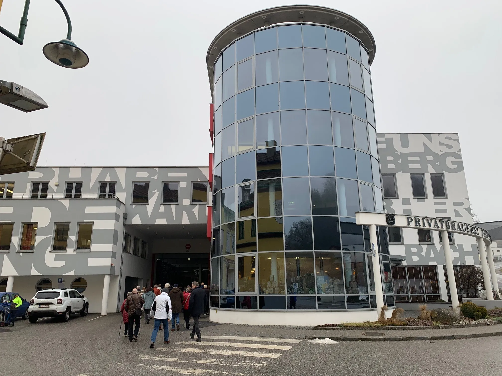 Eine Gruppe von Menschen geht an einem modernen Gebäude mit Glaswänden und einem zylindrischen Eingang vorbei. Das Gebäude hat ein Schild mit der Aufschrift 'UNBER'. Eine Straßenlaterne und ein weißer SUV sind in der Nähe.