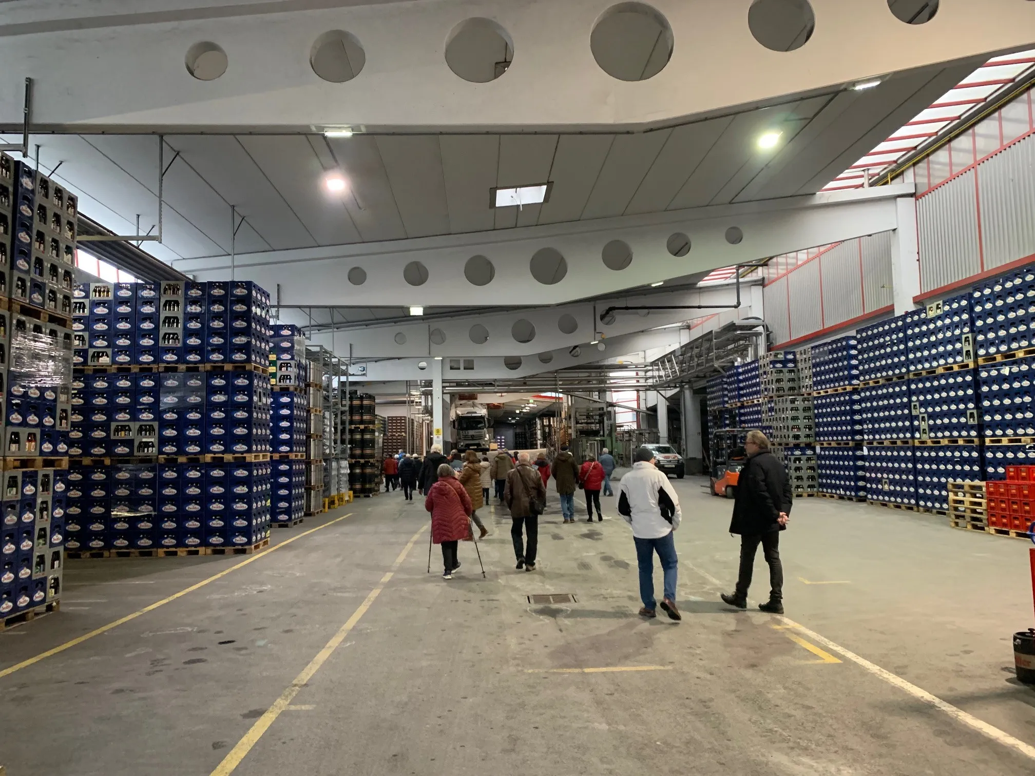 Ein Lagerhallen-Innenraum mit zahlreichen gestapelten Kisten und einigen Menschen, die gehen. Die Decke ist hoch mit runden Lichtern, und ein LKW ist geparkt.