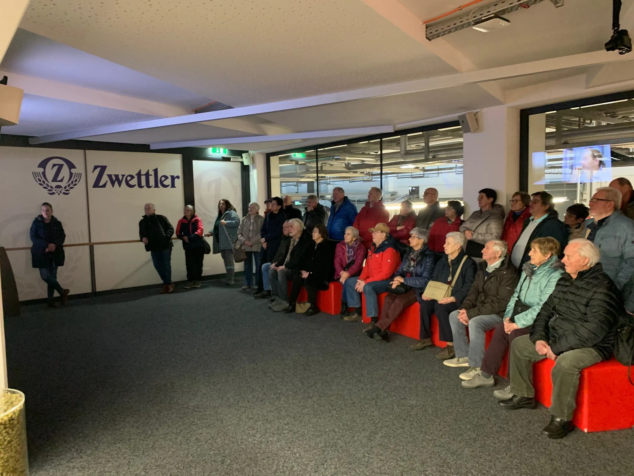 Eine Gruppe von Menschen sitzt auf roten Bänken in einem Raum mit Glaswänden, wahrscheinlich in Erwartung einer Veranstaltung. An der linken Wand steht das Wort 'Zwettler.' Einige Menschen stehen hinter der Wand.