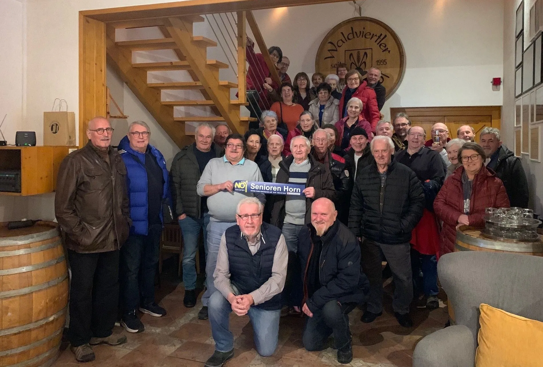 Eine Gruppe älterer Menschen versammelt sich für ein Foto in einem Raum mit einer Holztreppe. Ein Mann hält ein Schild mit der Aufschrift NOY Senioren Horn.