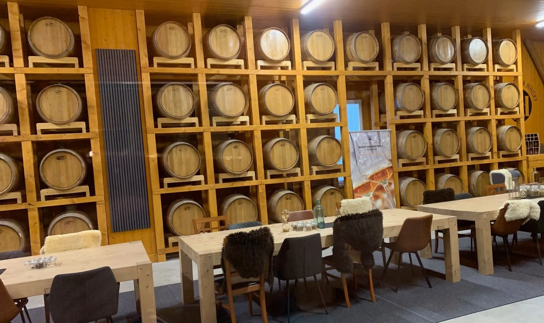 Ein Raum mit einem großen Holztisch, umgeben von Stühlen mit Fellbezug, einer Flasche und Weingläsern. Im Hintergrund sind Holzfässer auf Regalen gestapelt.