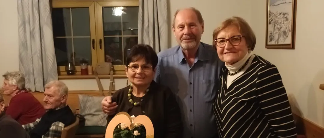 Drei Personen stehen lächelnd für die Kamera in einem Raum. Die Frau in der Mitte hält einen herzförmigen Rahmen mit Blumen. Hinter ihnen sitzen mehrere Personen um einen Tisch.