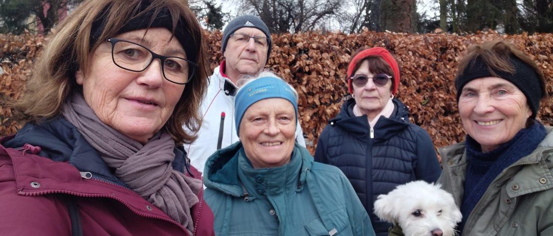 Fünf Personen und ein weißer Hund stehen vor einer Hecke und lächeln für ein Foto. Sie tragen Winterkleidung, und die Frau in der Mitte hat eine blaue Kopfband.