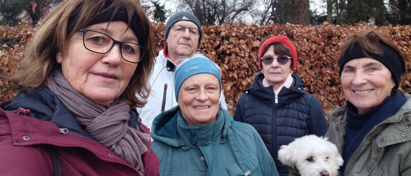 Fünf Personen und ein weißer Hund stehen vor einer Hecke und lächeln für ein Foto. Sie tragen Winterkleidung, und die Frau in der Mitte hat eine blaue Kopfband.