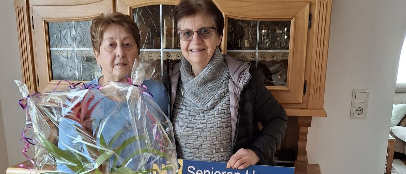 Zwei Frauen stehen in einem Raum und halten eine Topfpflanze. Eine hält ein blaues Schild mit 'Senioren Horn' darauf. Hinter ihnen steht ein Holzschrank mit Glastüren.