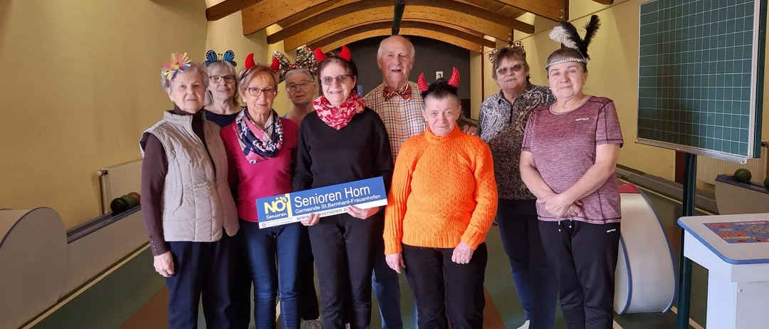 Eine Gruppe Senioren steht in einer Bowlingbahn. Sie sind alle in festlicher Kleidung gekleidet und halten ein blaues Schild mit der Aufschrift 'Senioren Horn'.