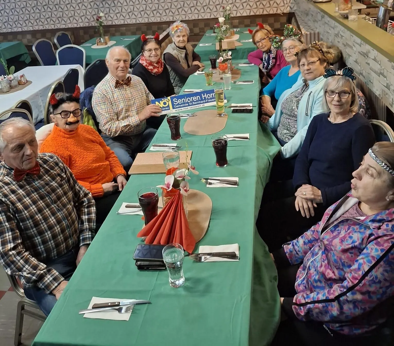 Eine Gruppe älterer Menschen mit festlichen Hörnern sitzt in einem Restaurant um einen Tisch herum, mit verschiedenen Getränken und einem Banner, das NO! Senioren Horn steht.