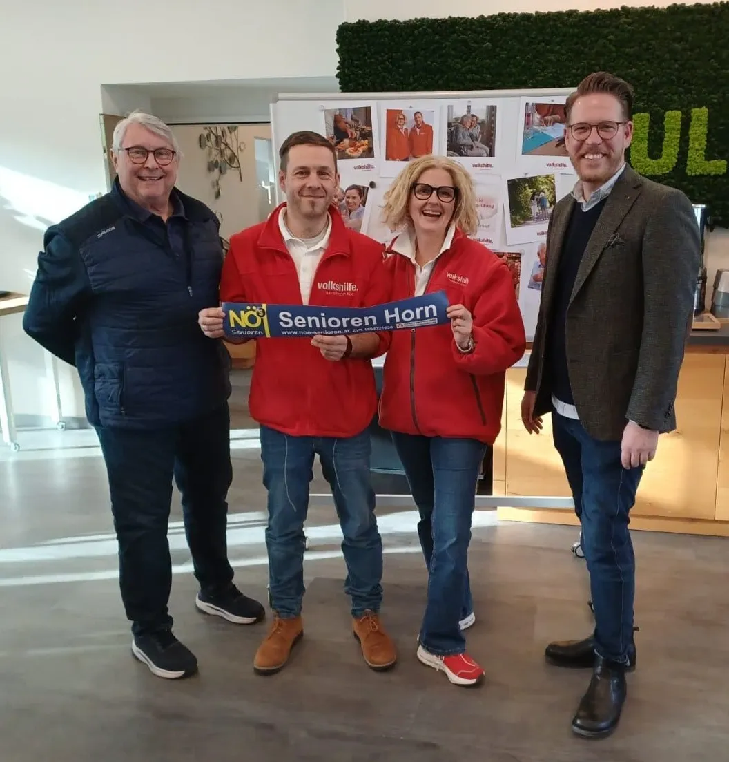 Vier Menschen posieren für ein Foto, zwei halten ein blaues Schild mit der Aufschrift 'Senioren Horn'. Die beiden in roten Jacken stehen in der Mitte. Dahinter befindet sich eine Wand mit Fotos und einem großen Buchstaben 'U'.