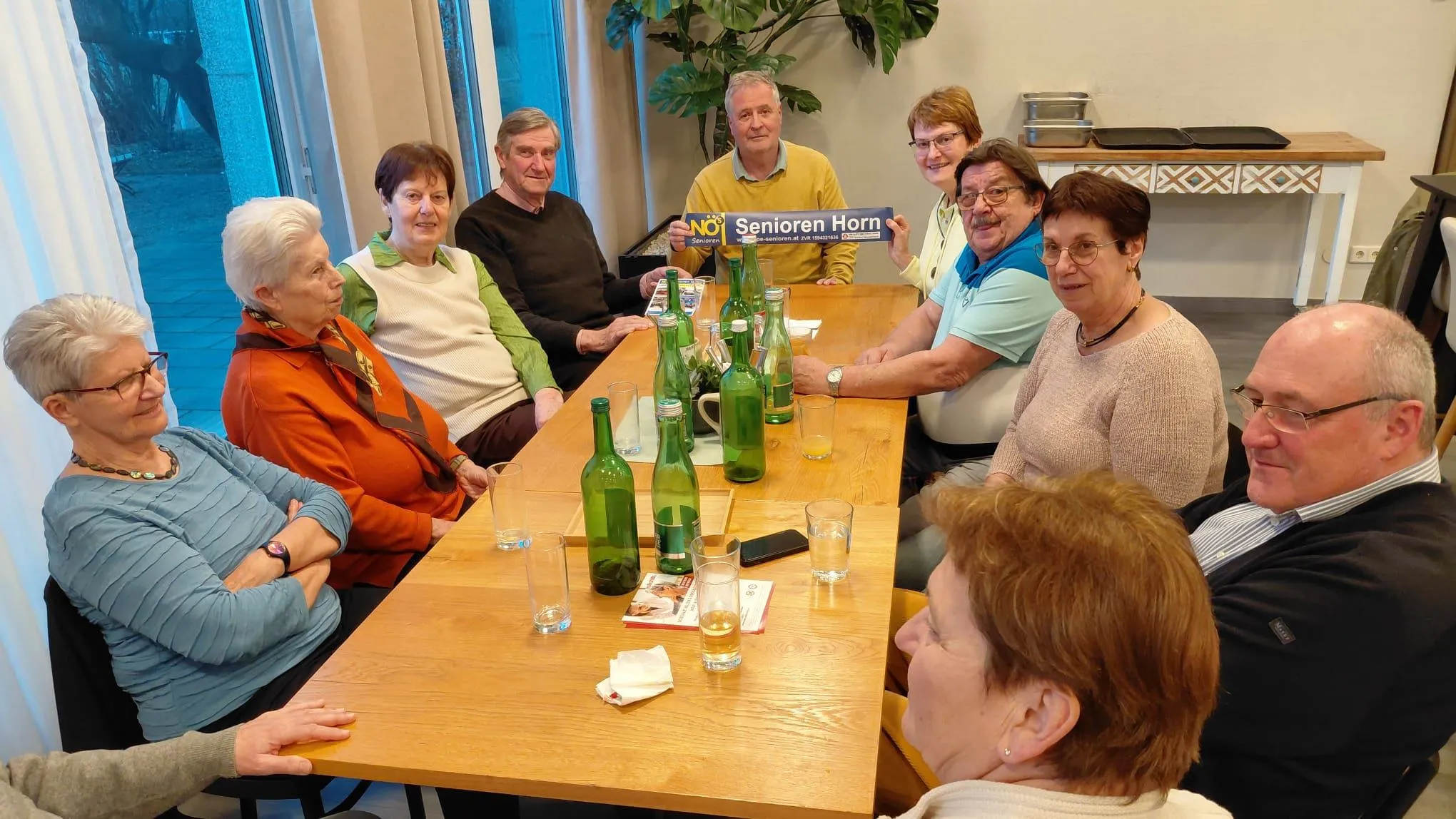 Eine Gruppe älterer Menschen sitzt um einen Tisch herum, lächelt und posiert für ein Foto. Ein Mann hält ein blaues Banner, auf dem NO Senioren Horn steht.