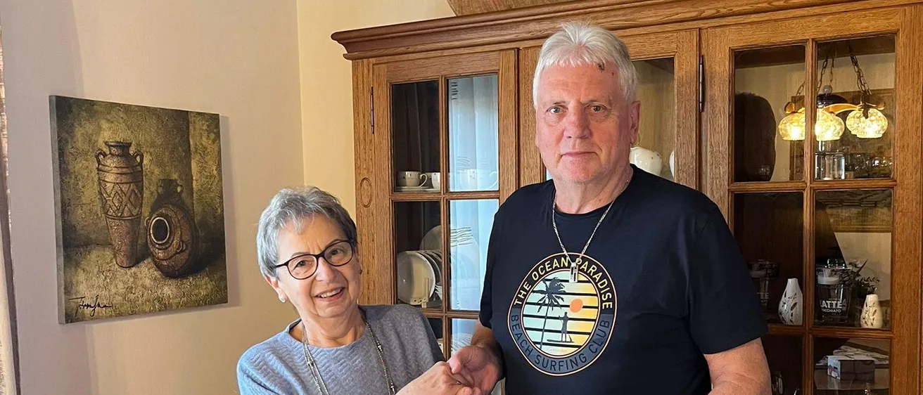 Eine ältere Frau mit Brille übergibt einem Mann in einem schwarzen T-Shirt ein Geschenk in einem Wohnzimmer mit einem Holzschrank und einem Gemälde an der Wand.