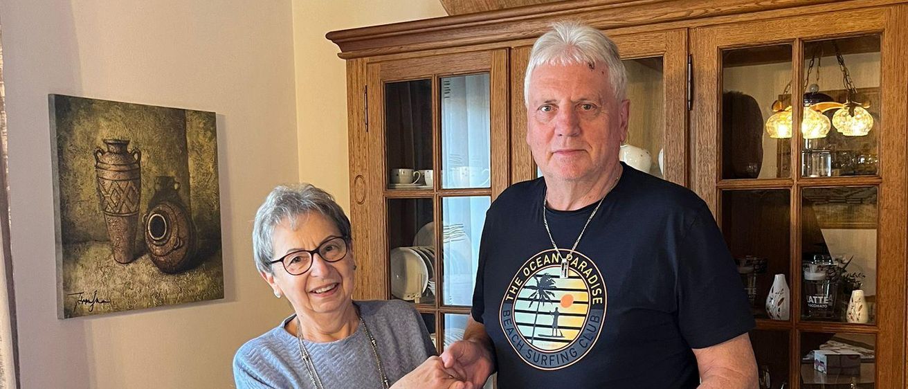 Eine ältere Frau mit Brille übergibt einem Mann in einem schwarzen T-Shirt ein Geschenk in einem Wohnzimmer mit einem Holzschrank und einem Gemälde an der Wand.