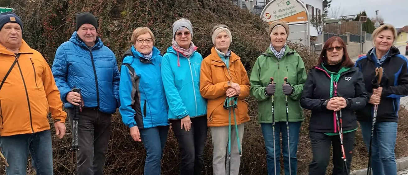 Eine Gruppe von sieben Menschen in warmen Kleidungsstücken und mit Wanderstöcken posiert für ein Foto vor einer Hecke.