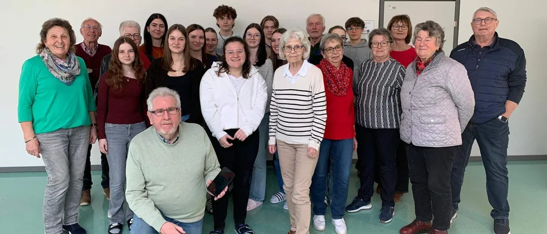Eine Gruppe von Menschen in einem Raum, einige stehen und einige knien, lächeln für ein Foto. Ein Mann hält ein Smartphone. Der Raum hat weiße Wände und grünen Boden.