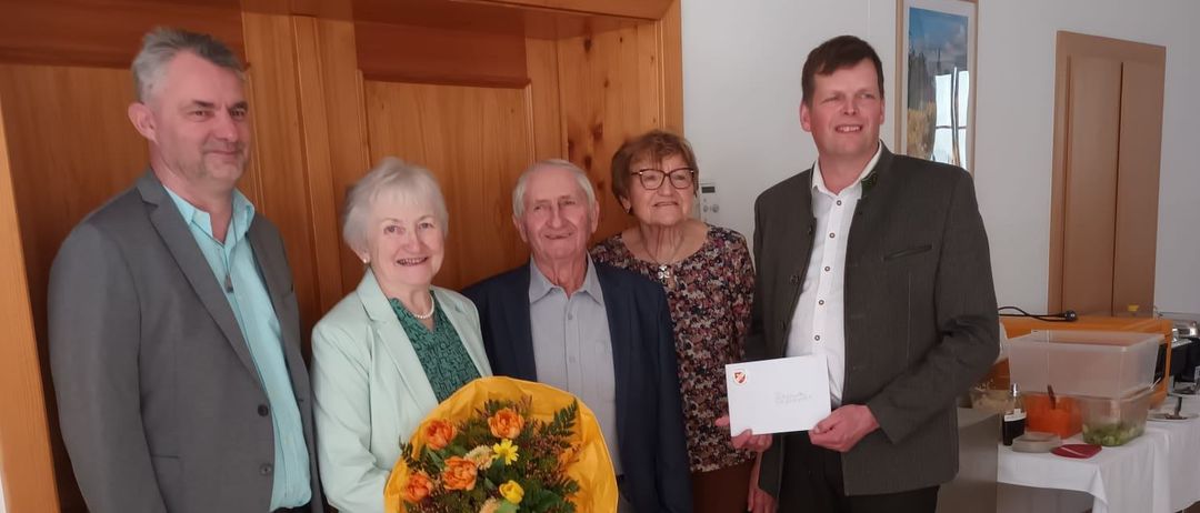 Fünf Personen stehen in einem Raum mit Holzwänden. Ein Mann hält ein Papier und eine Frau Blumen. Sie scheinen einen Anlass zu feiern. Dahinter steht ein Tisch mit Essen.