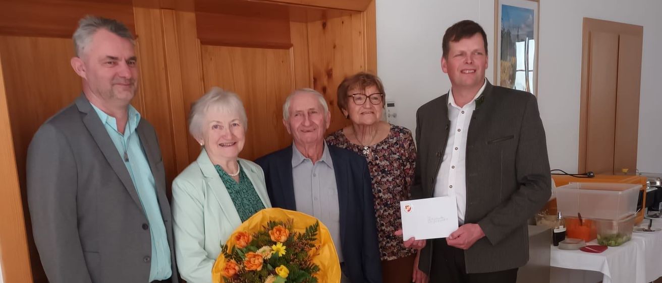 Fünf Personen stehen in einem Raum mit Holzwänden. Ein Mann hält ein Papier und eine Frau Blumen. Sie scheinen einen Anlass zu feiern. Dahinter steht ein Tisch mit Essen.
