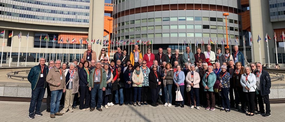 Eine Gruppe von Menschen in Jacken und Turnschuhen posiert für ein Foto vor einem modernen Gebäude mit Flaggen und klarem Himmel.