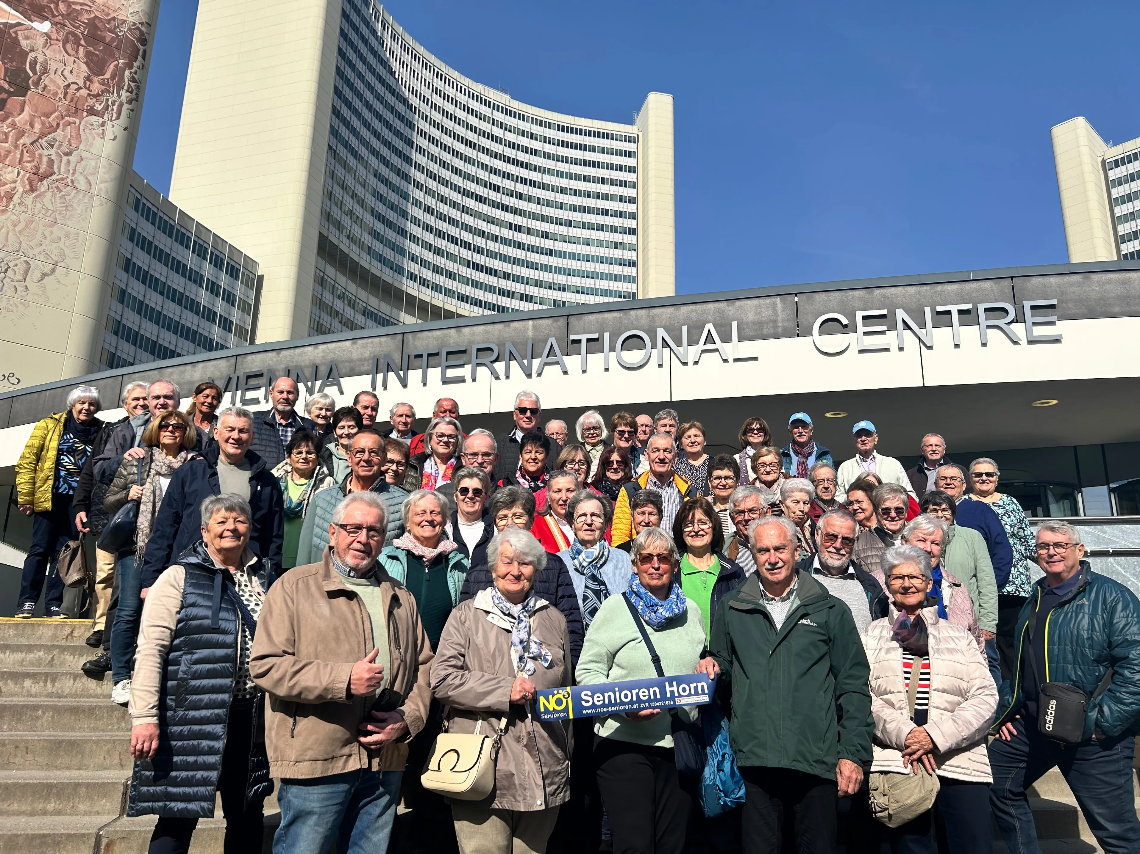 Eine Gruppe älterer Menschen posiert für ein Foto vor dem Wiener Internationalen Zentrum, einer hält ein blaues Schild mit der Aufschrift 'Senioren Horn'.