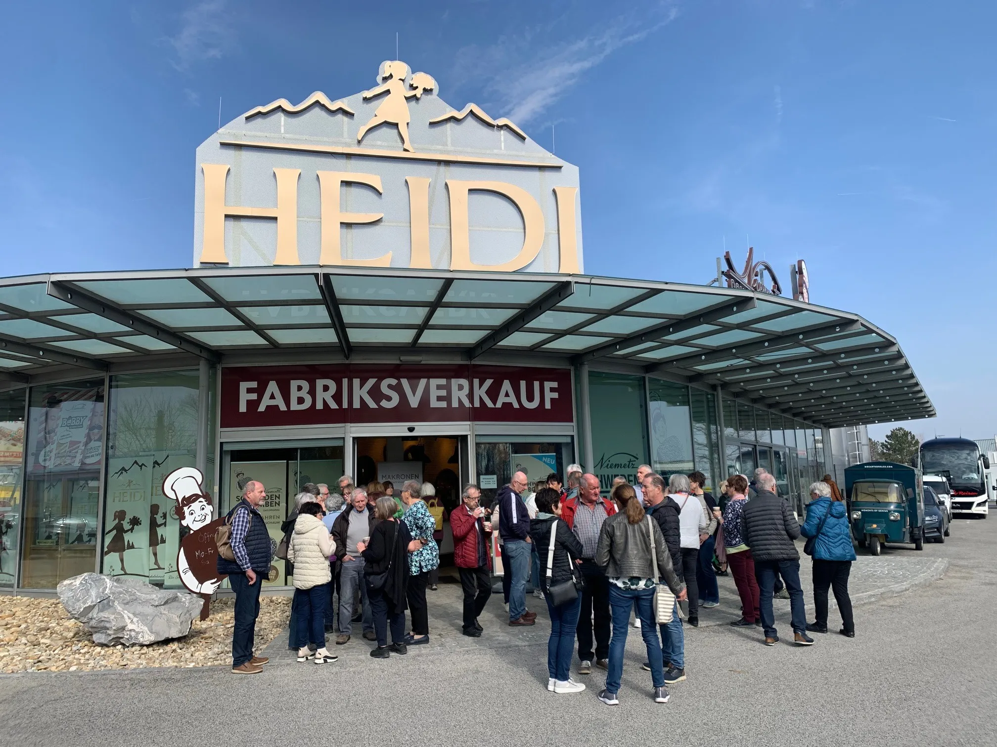 Eine Gruppe von Menschen steht vor dem Heidi's Factory Store, einem Gebäude mit Glaswänden und einem Dach. Der Eingang ist mit einem Schild gekennzeichnet, das 'FABRIKSVVERKAUF' steht. Die Menschen sind in Winterkleidung gekleidet.